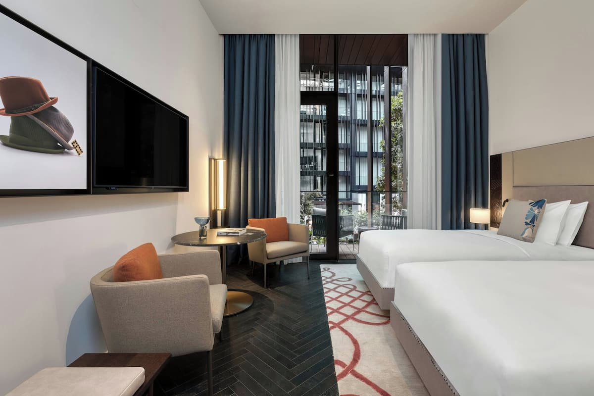 A hotel room with a bed, two armchairs, a small table, a TV mounted on the wall, and a view of the outside through large windows.