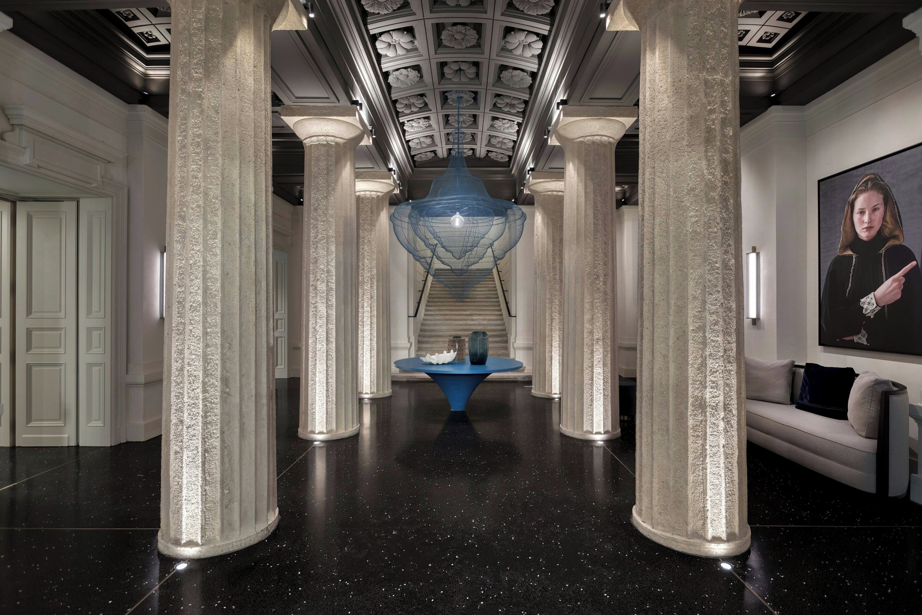 A grand hallway with high ceilings, ornate columns, and a central blue chandelier.
