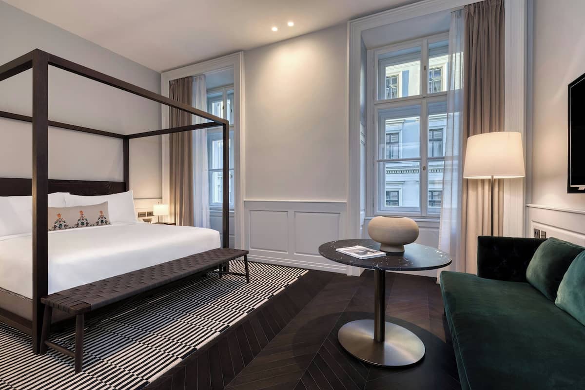 A modern hotel room with a four-poster bed, a round table with a lamp, a green sofa, and large windows with curtains.