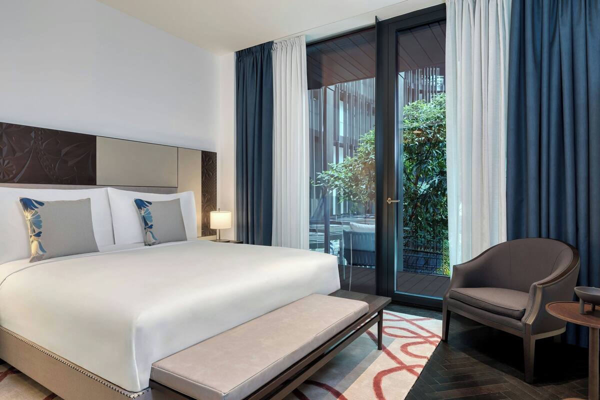 A hotel room with a large bed, a sofa, a small table, and a view of greenery through the window.
