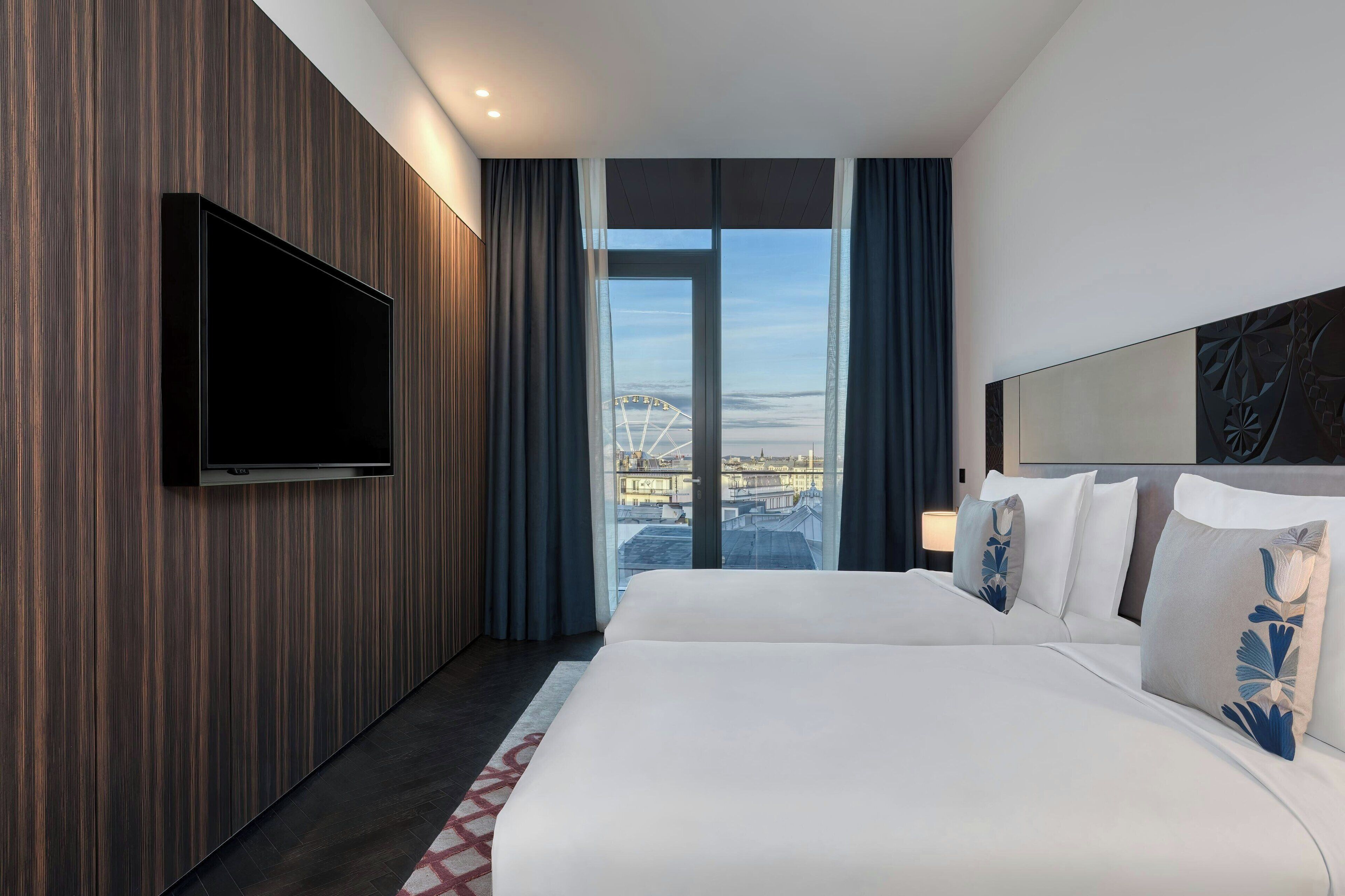 A hotel room with a large bed, a flat-screen TV mounted on the wall, and a view of a cityscape through a large window.