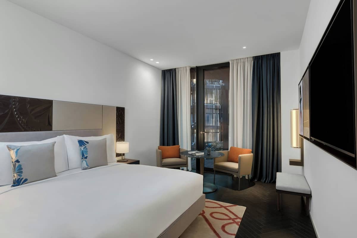 A modern hotel room with a large bed, two armchairs, a small table, and a TV.