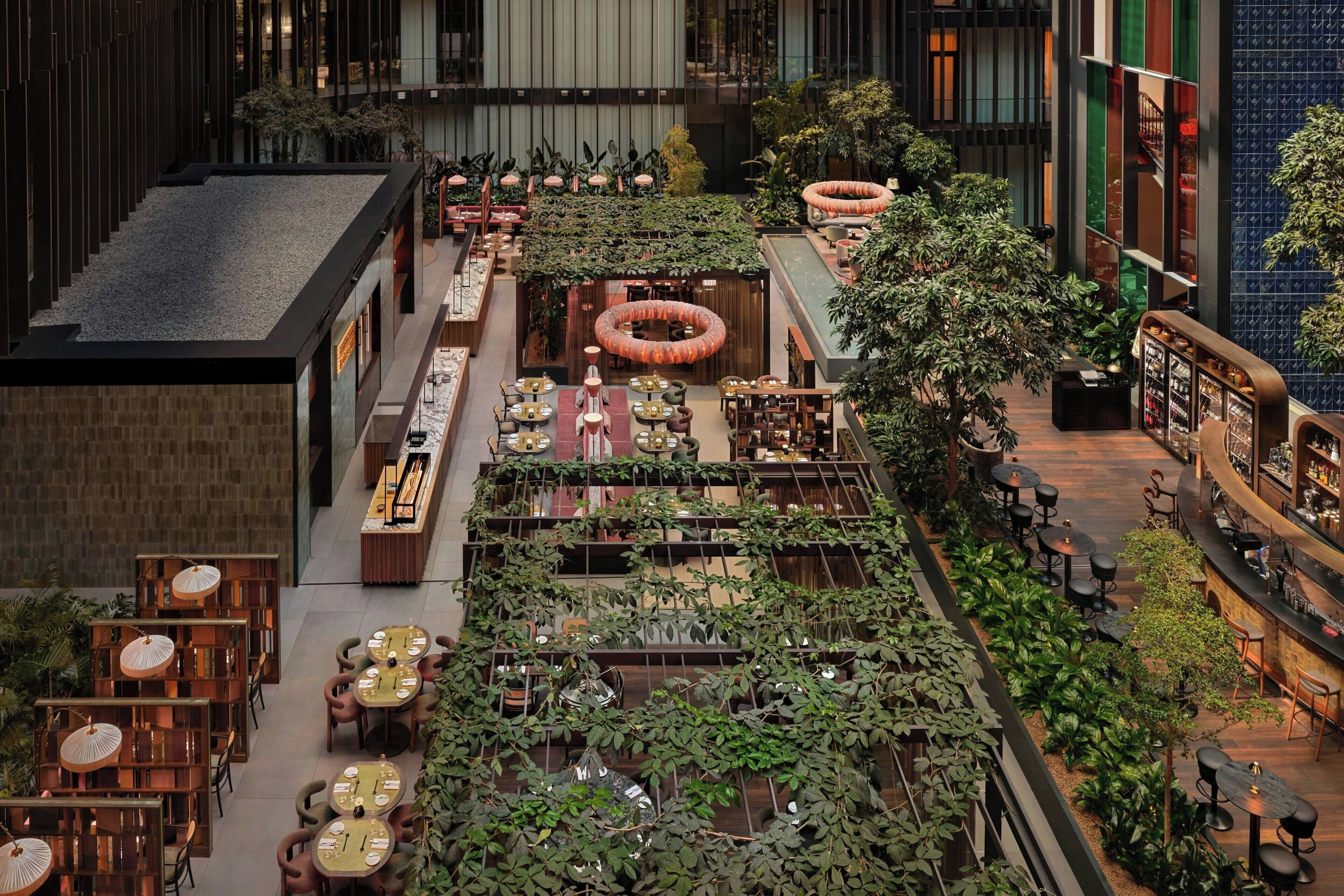 An indoor dining area with multiple tables and chairs, surrounded by greenery and a central water feature.