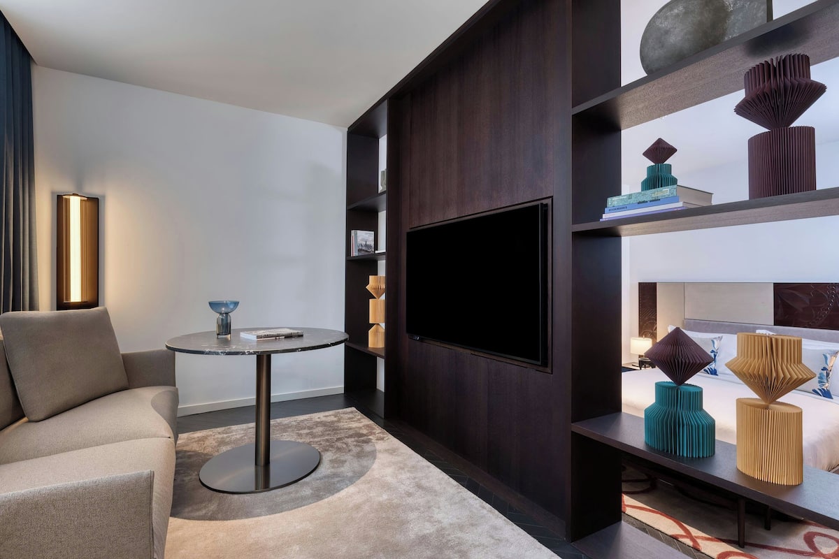 A modern living room with a sofa, a round coffee table, a flat-screen TV mounted on a dark wooden wall, and decorative vases on shelves.