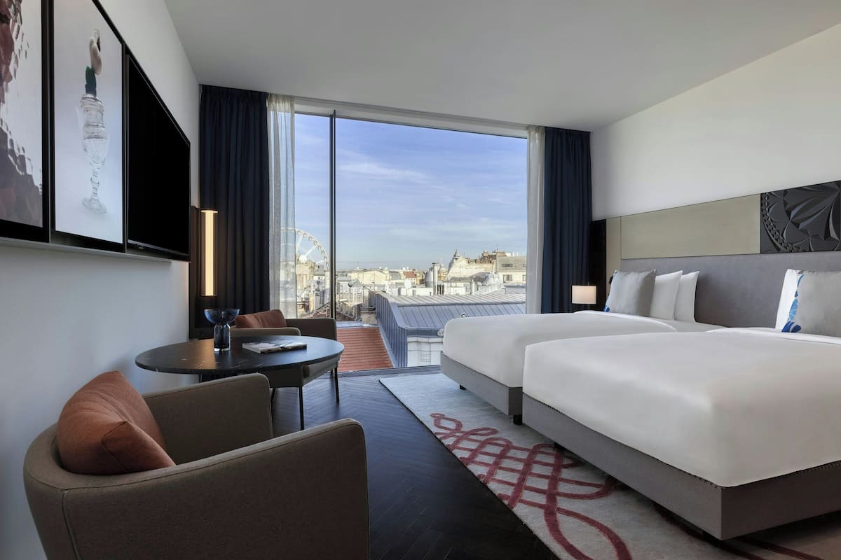 A hotel room with a large bed, a sofa, a small table, and a view of the cityscape through large windows.