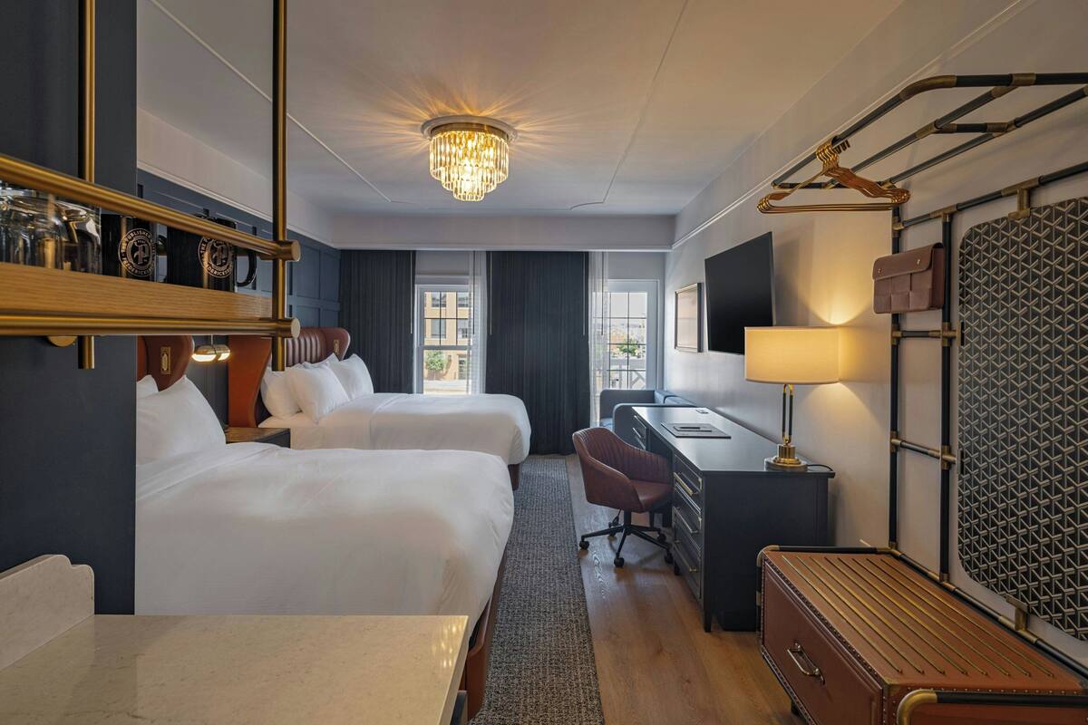 Ett hotellrum med två sängar, ett skrivbord, en stol, en resväska, en lampa, en spegel och en kristallkrona.