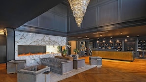 Un hall d’entrée spacieux avec une cheminée en marbre, des sièges recouverts d’un tissu gris, un plancher en bois et un coin bar avec des étagères.