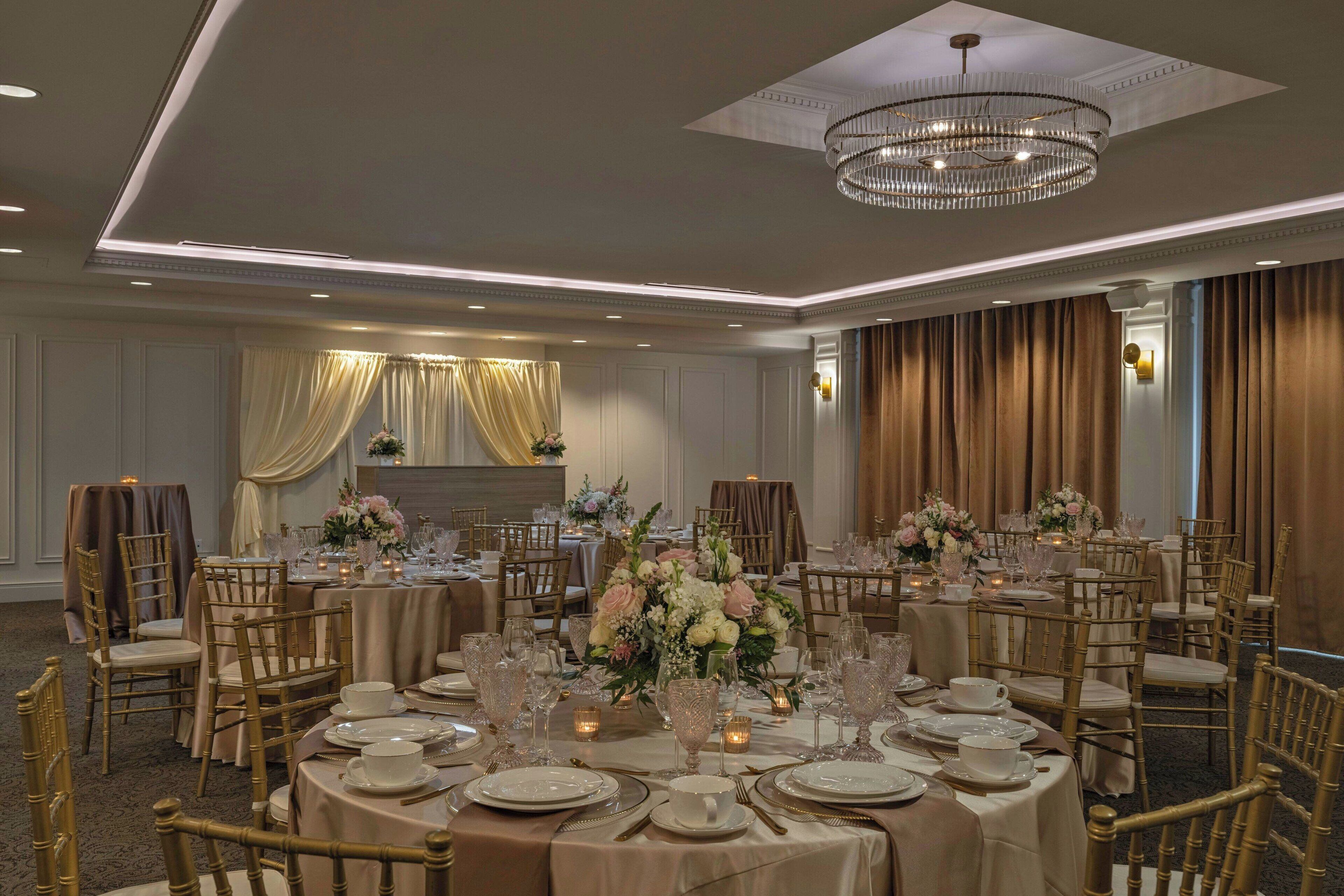 A banquet hall with round tables set for dining, gold chairs, and a chandelier.