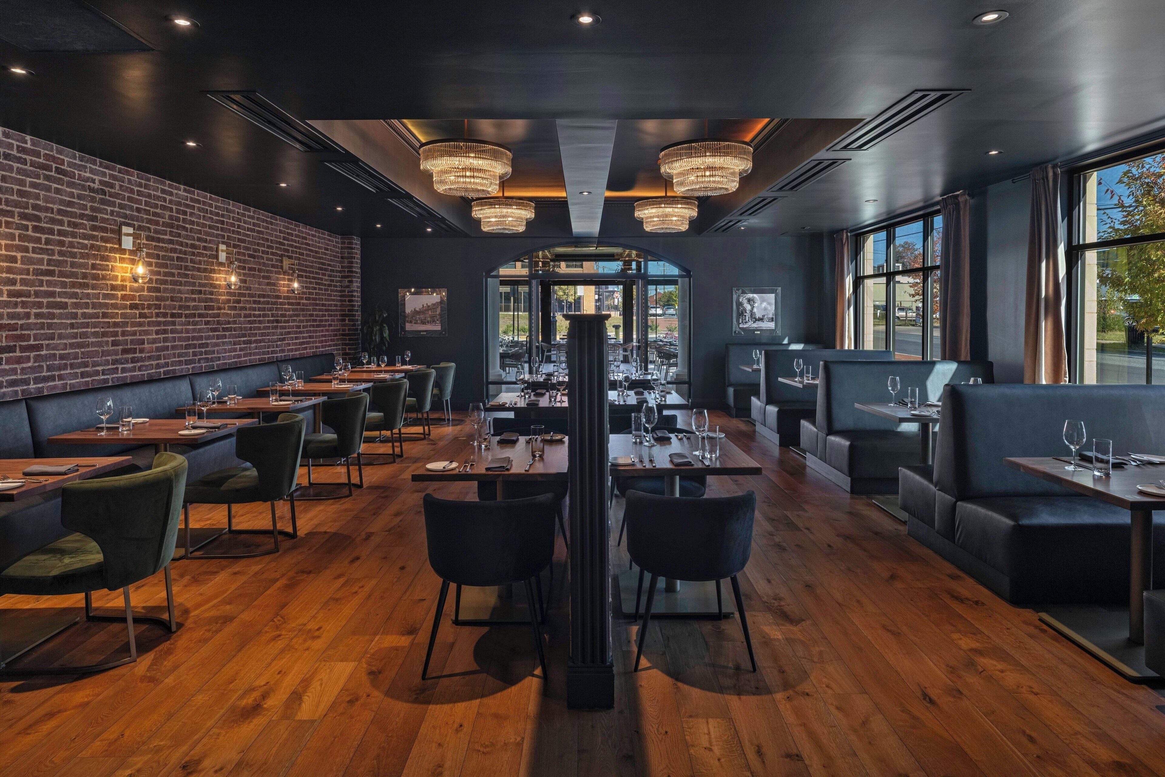 A restaurant with a brick wall, wooden flooring, and modern chandeliers. Tables are set with chairs and dining ware.
