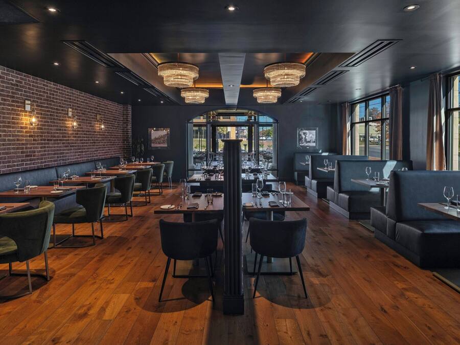 A restaurant with a brick wall, wooden flooring, and modern chandeliers. Tables are set with chairs and dining ware.