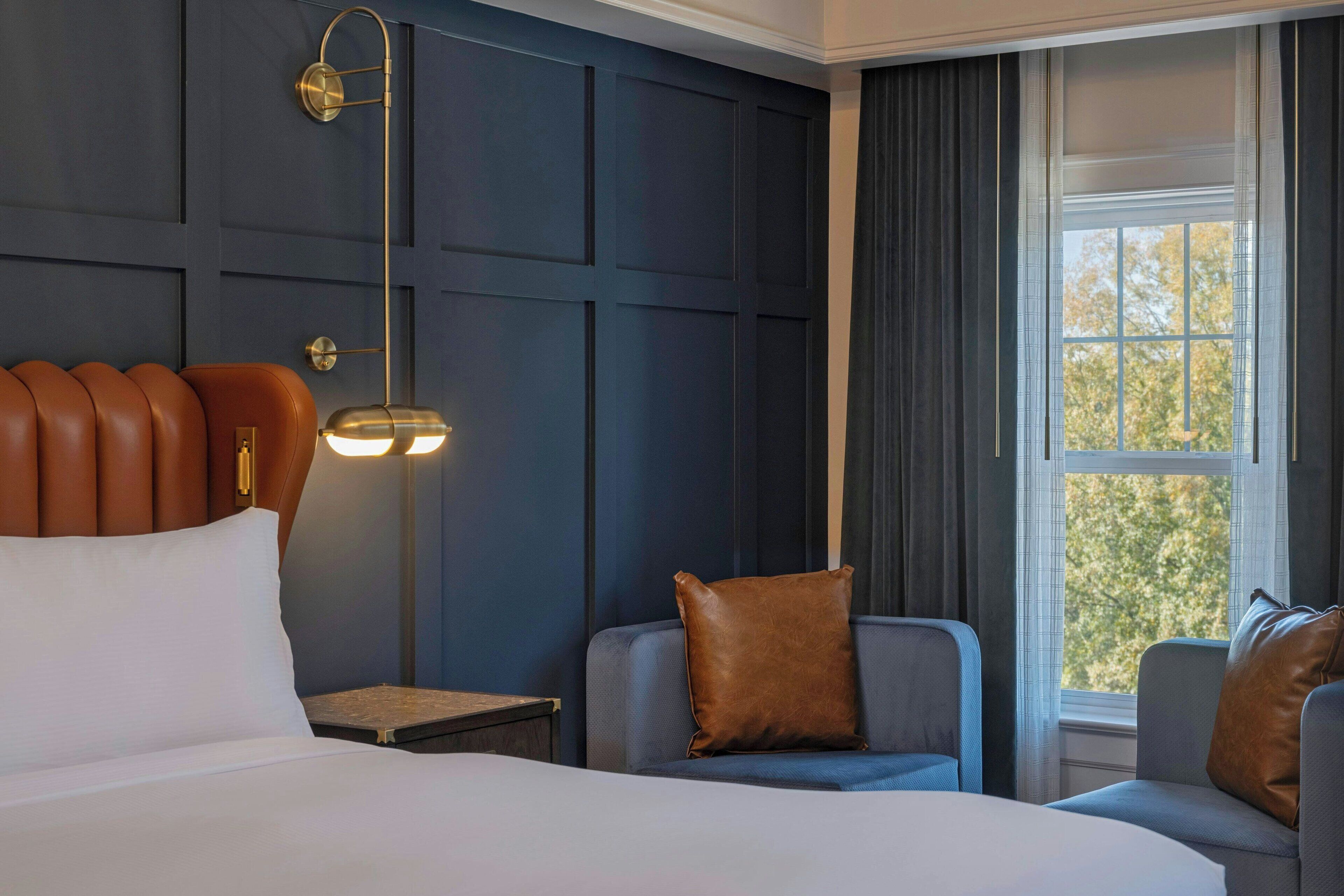 A hotel room with a bed, two armchairs, a nightstand, a lamp, and a window with curtains.