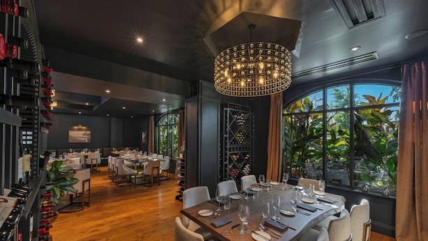 Un restaurant avec une grande table dressée pour un repas, entourée de chaises. On aperçoit un meuble à vin avec des bouteilles sur le côté gauche, et un lustre suspendu au plafond.
