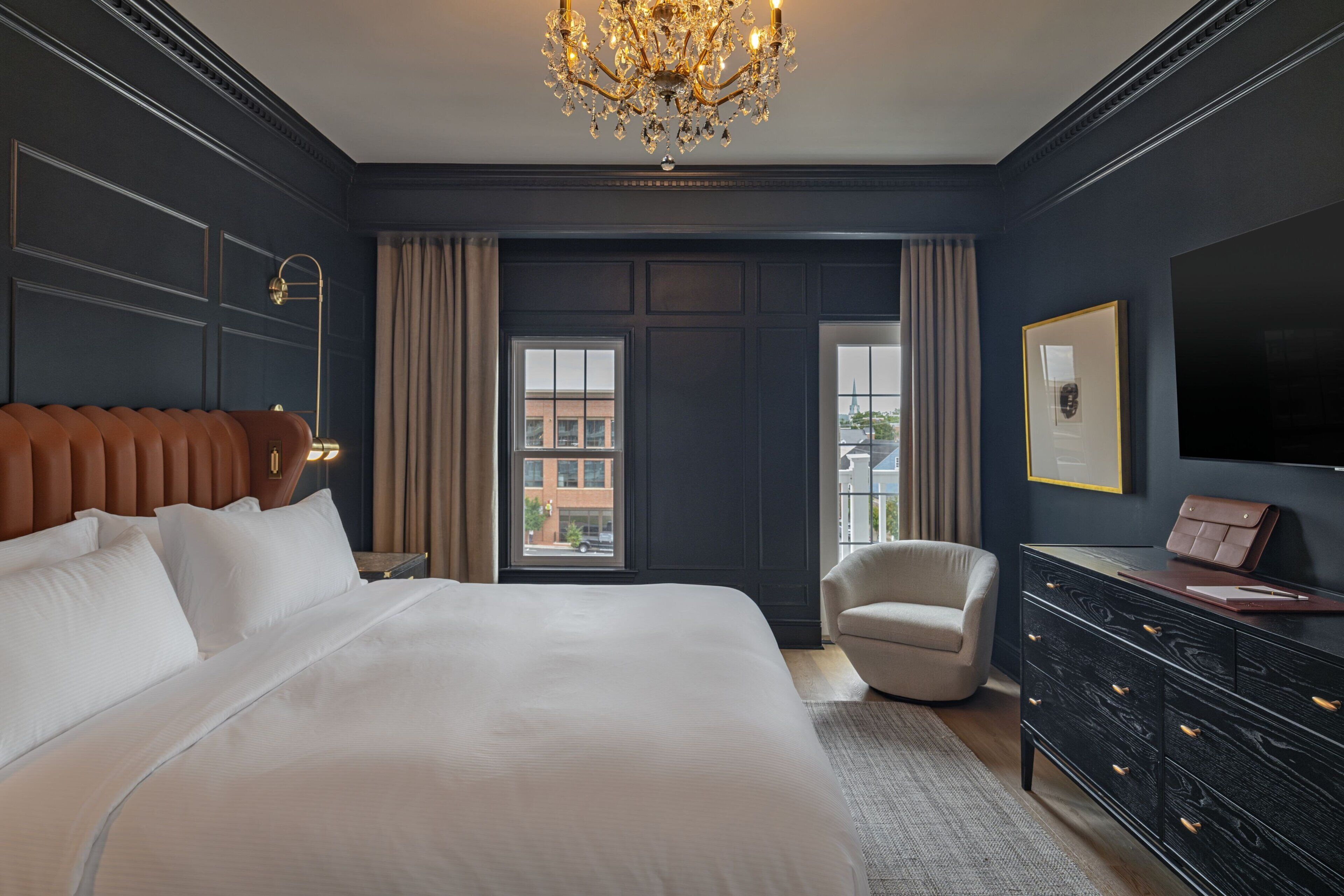 Un dormitorio amplio con una cama grande, un candelabro, televisión y vistas de la ciudad a través de la ventana.