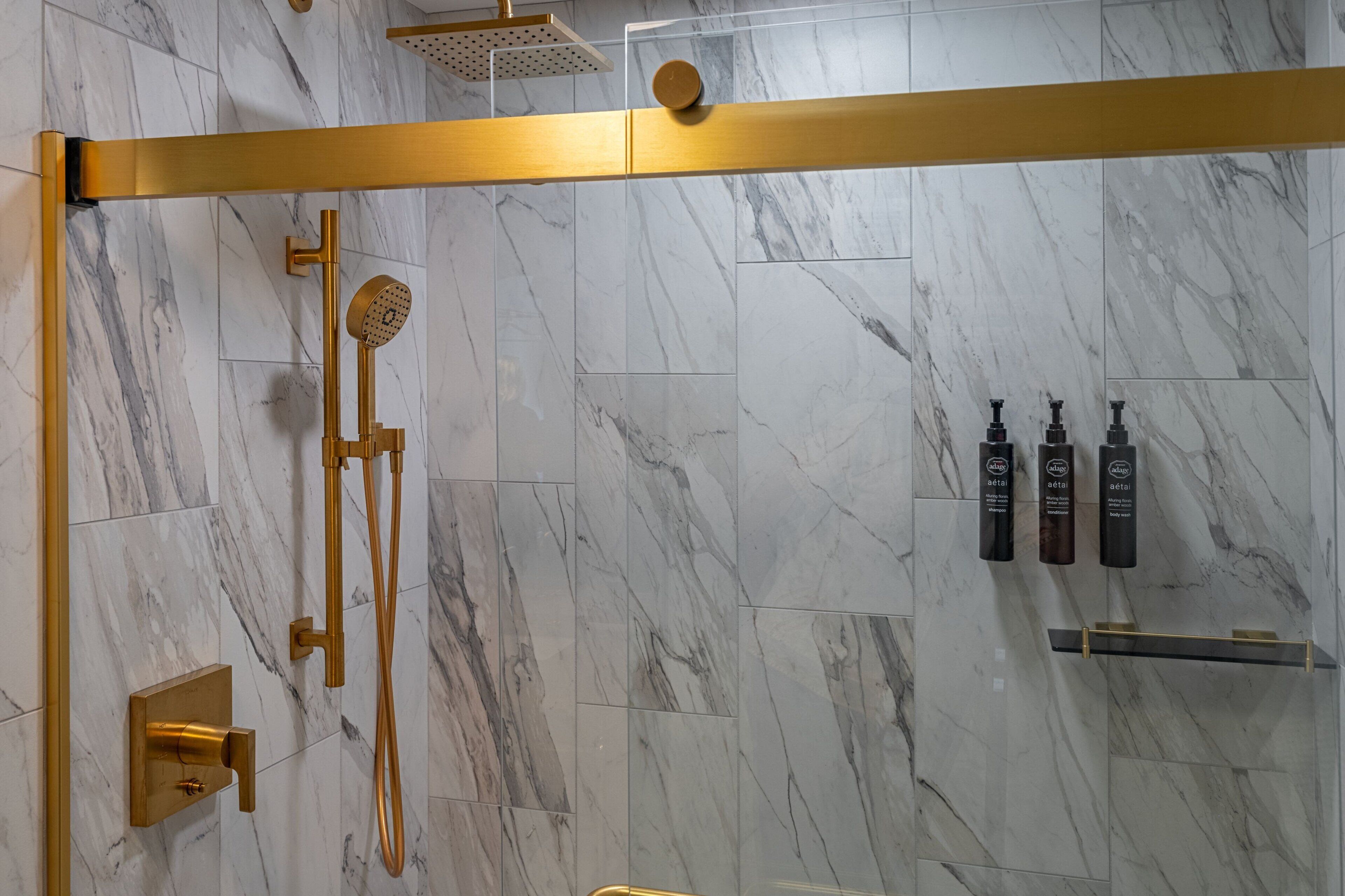 Une salle de bain moderne avec des murs en marbre, une cabine de douche dorée et trois bouteilles de shampoing.