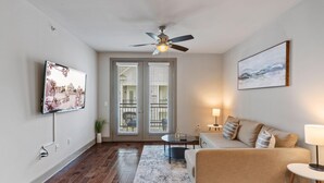 A modern living room with a sofa, a coffee table, a TV mounted on the wall, a ceiling fan, and a large window with blinds. - VA - Sutton - Apt 5119 (Woodbridge)