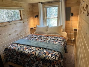 A wooden cabin bedroom with a bed, two windows, a nightstand, and a wall-mounted lamp. - Eve's Place Lakeside Cabin 4: Pet Friendly! (Grand Marais)