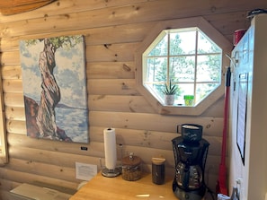 Interior de uma cabana de madeira com um quadro de uma árvore, uma janela com vista para árvores, uma cafeteira e um dispensador de toalhas de papel.