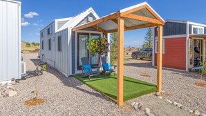 Une maison mobile dotée d’un patio couvert, de deux chaises bleues et d’un pot de fleurs suspendu.