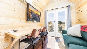 A cozy cabin interior with a wooden desk, two chairs, a television, a couch, and a sliding glass door leading to an outdoor area. - Luxury Tiny Home/Base Camp to Ouray, Telluride/2 Queen beds, 1 twin bed/Washer & Dryer/Smart TV/Hot Tub Access/Patio w/Mountain Views/ Fire Pit/11 mi to Airport (Montrose)