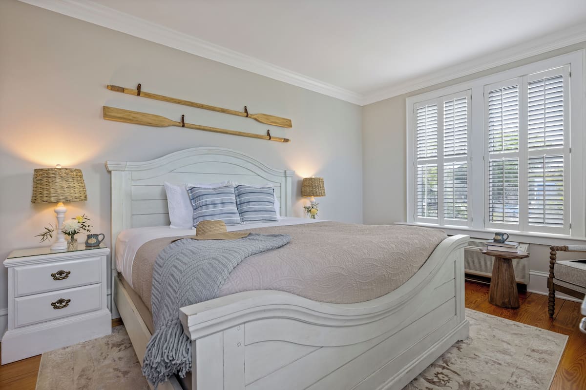 A bedroom with a white bed, two bedside tables, a nightstand, a window with blinds, and a small wooden stool.