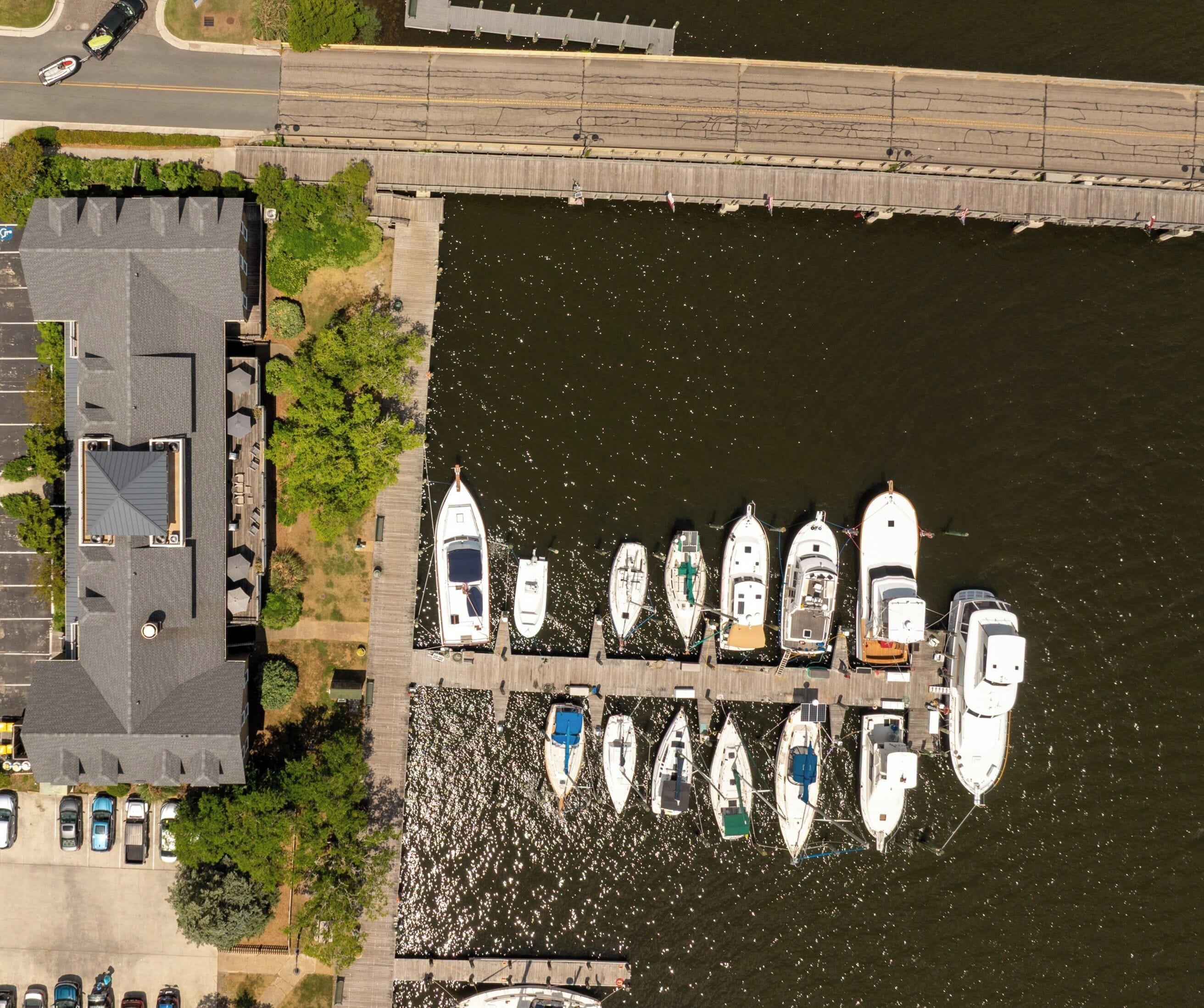 An aerial view of a marina with boats docked in a row, a parking area with cars, and a building with a sloped roof.
