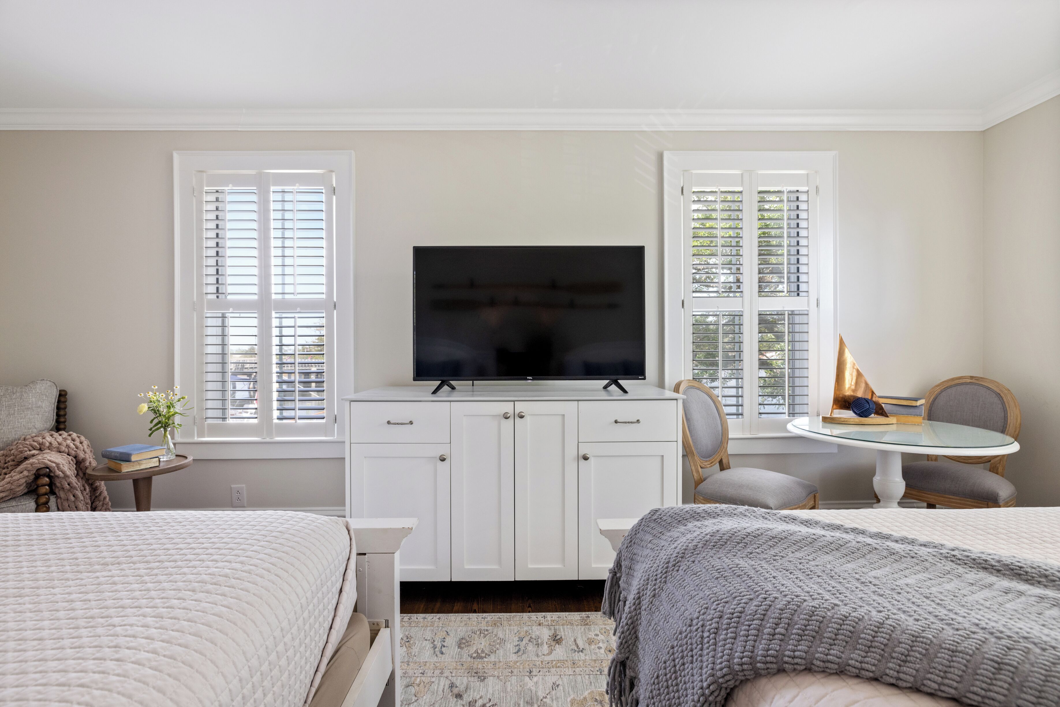 A bedroom with a bed, a desk with a television, a chair, and a small table with books and a vase.