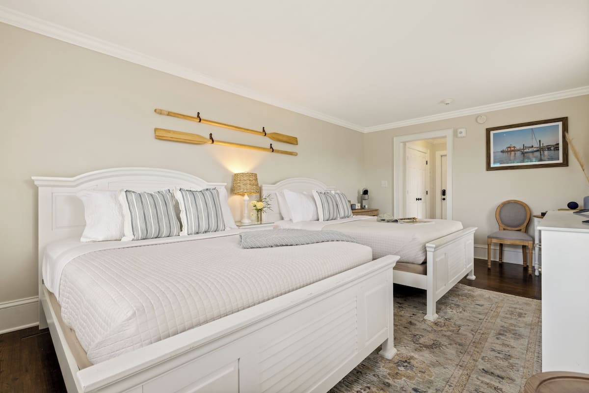 A bedroom with two white beds, a wooden headboard, a lamp, and a painting on the wall.