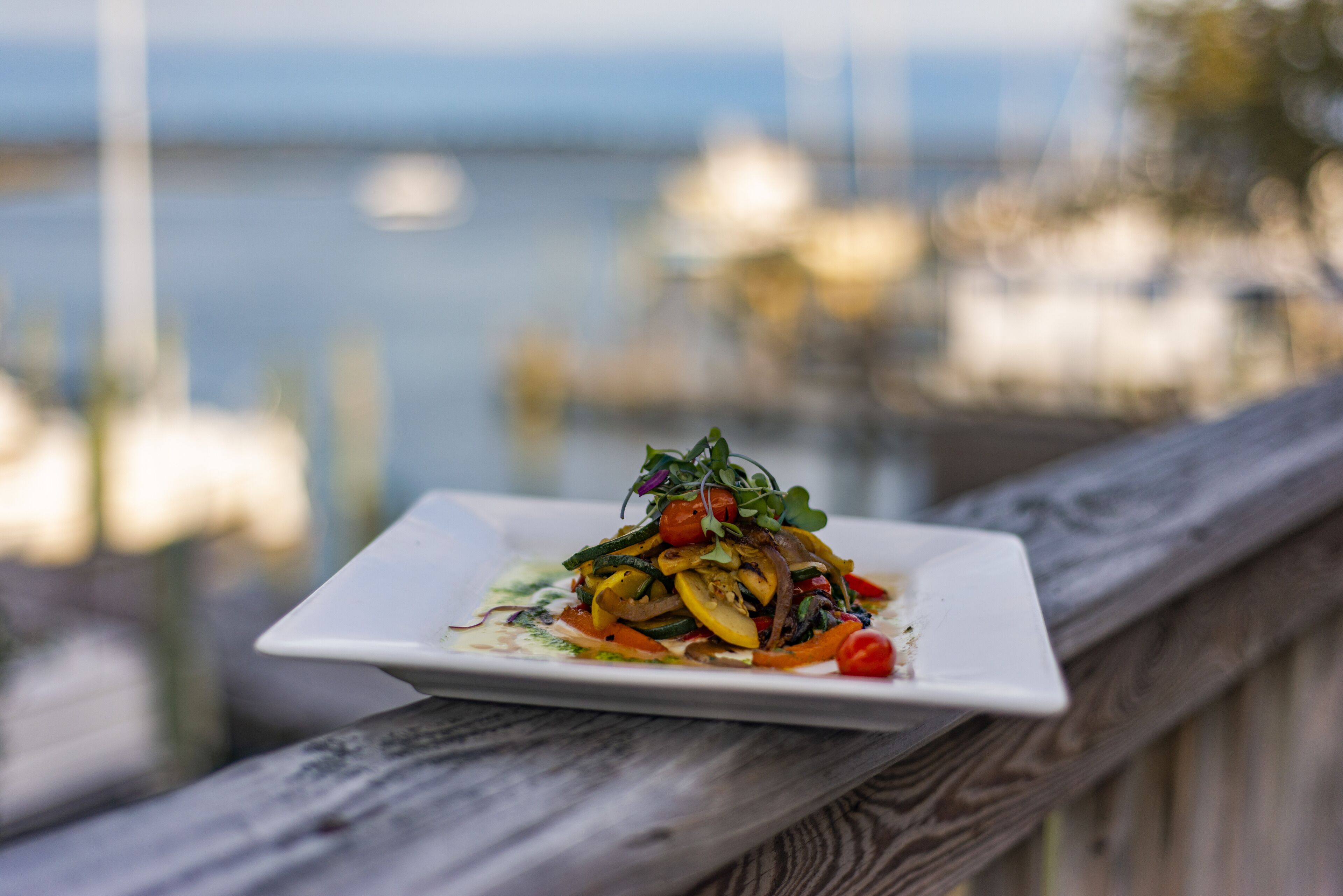 A square white plate with a colorful vegetable dish on a wooden railing overlooking a body of water.