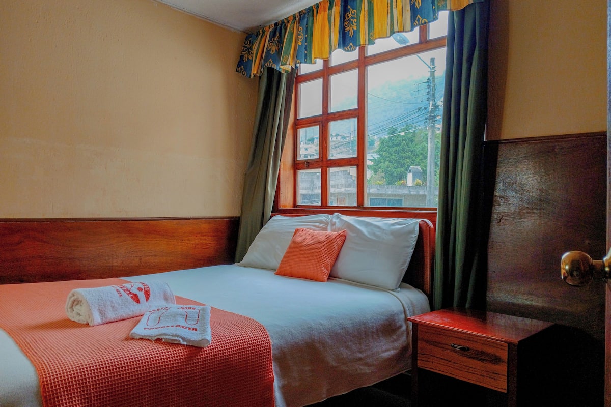 Une chambre d’hôtel avec un lit, une table de chevet en bois, une fenêtre avec des rideaux et une vue sur l’extérieur.