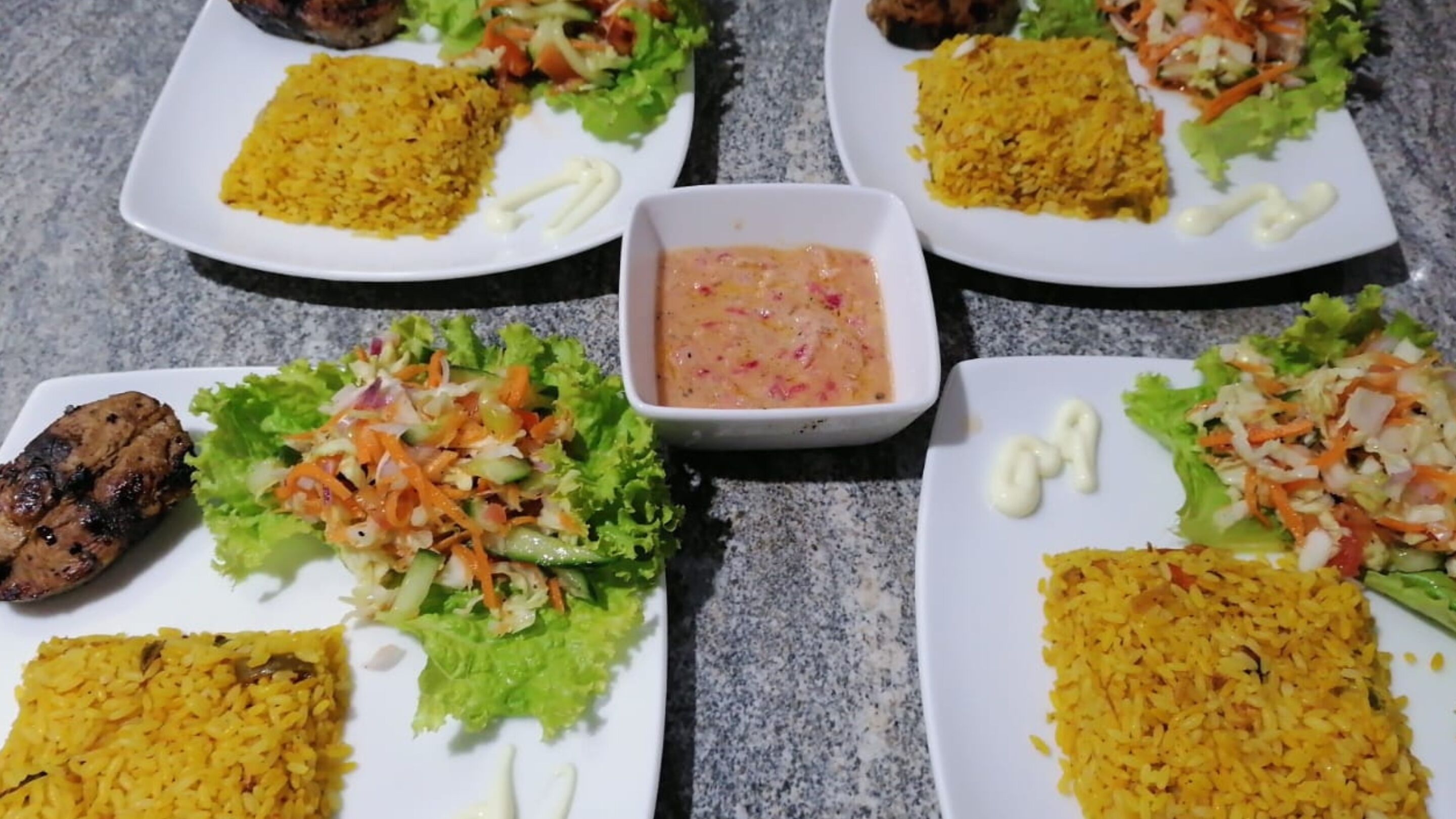 Quatre assiettes garnies de viande grillée, de riz jaune et d’une salade, accompagnées d’un petit bol de sauce.