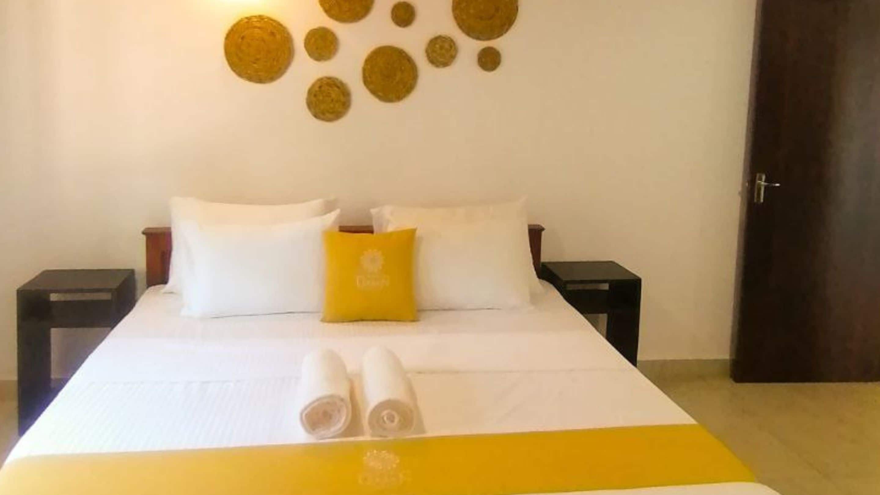 A neatly made bed with white and yellow bedding, two white pillows, and a yellow accent pillow. There are two bedside tables with lamps, and a door is visible to the right.