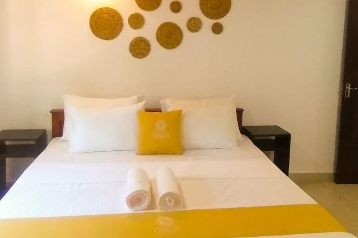 A neatly made bed with white and yellow bedding, two white pillows, and a yellow accent pillow. There are two bedside tables with lamps, and a door is visible to the right.