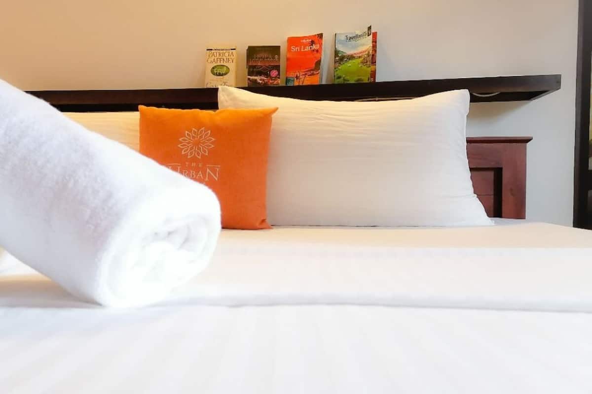 Una cama bien hecha con sábanas blancas y una toalla blanca enrollada. Hay un almohadón naranja con diseño floral y una almohada blanca. Un estante sobre la cama contiene varios libros de viajes.