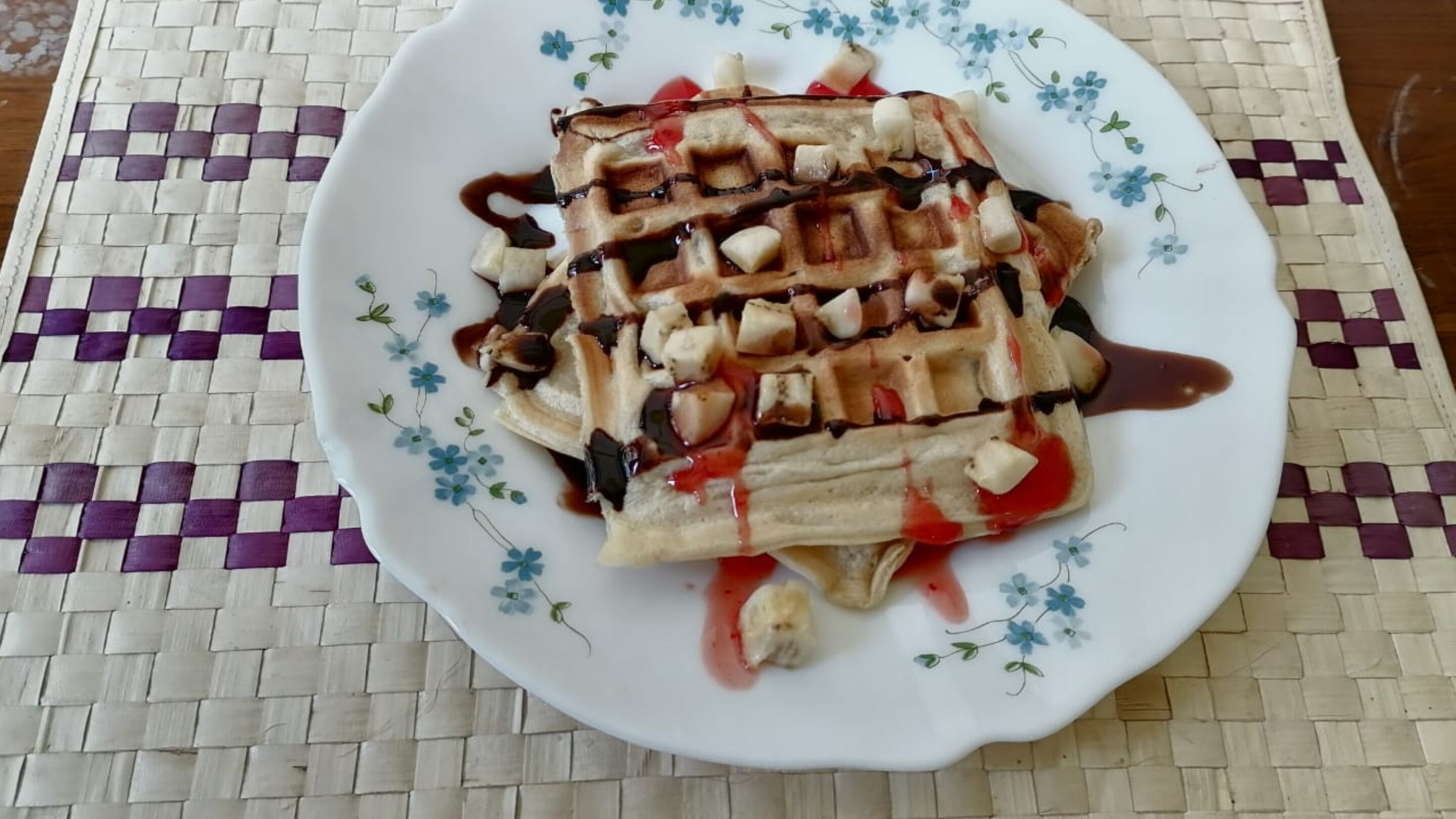 A plate of waffles with chocolate sauce and fruit on a patterned tablecloth.