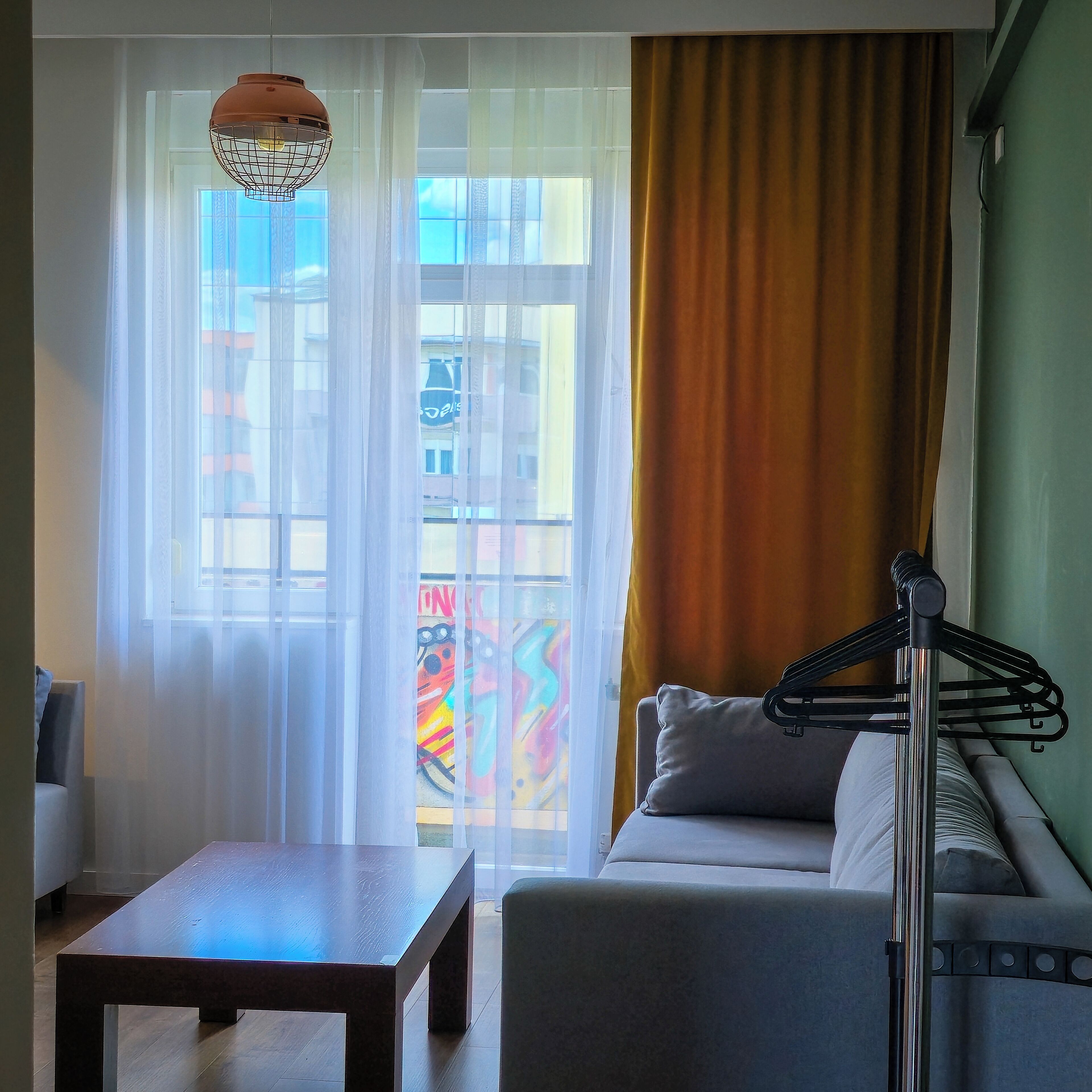 Una sala de estar con sofá, mesa de centro, lámpara colgante, cortinas translúcidas y vistas de edificios a través de una ventana.