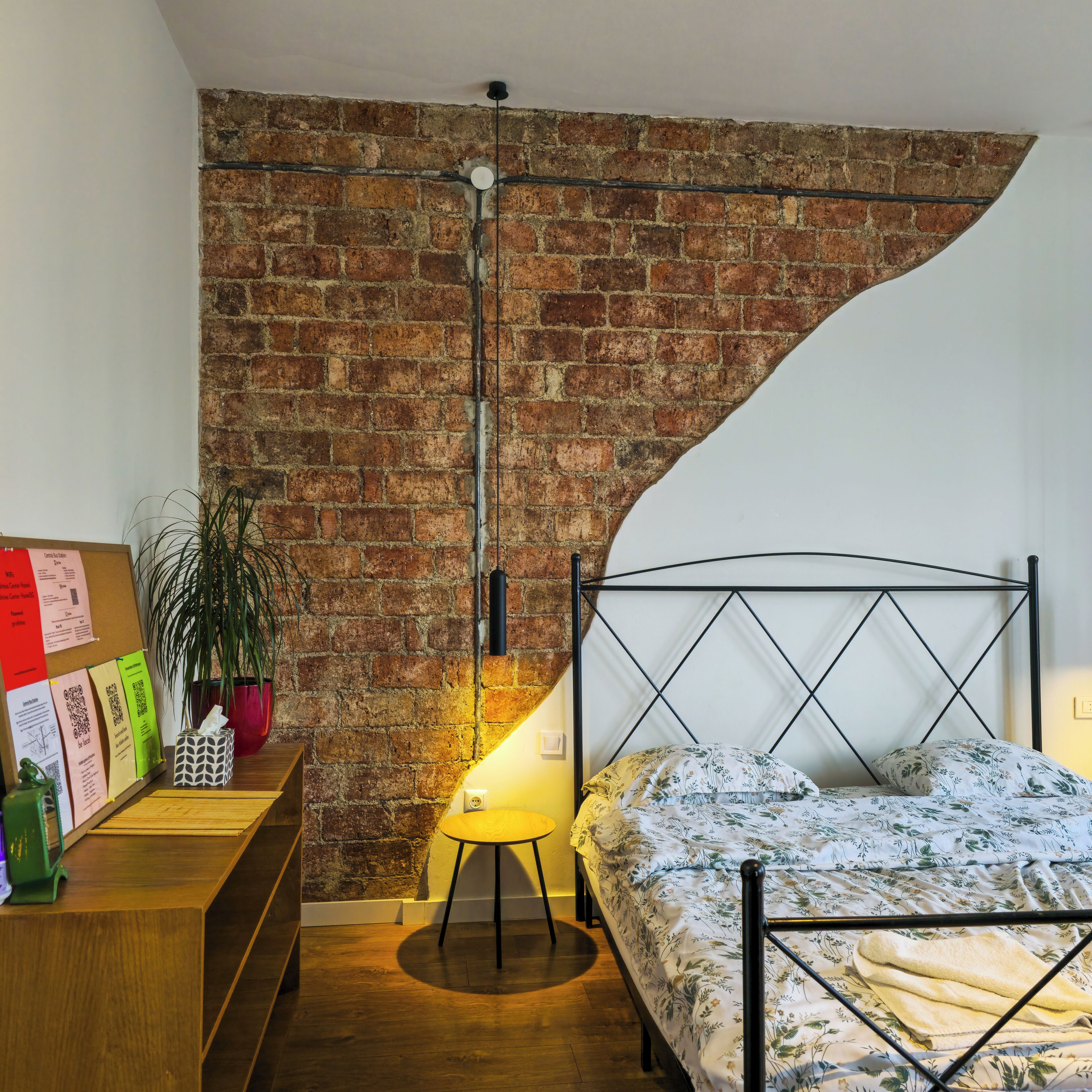 Une chambre avec un mur en briques, un lit recouvert d’une couvre-lit à motifs floraux, une lampe sur pied et une table de chevet en bois sur laquelle est posée une plante.