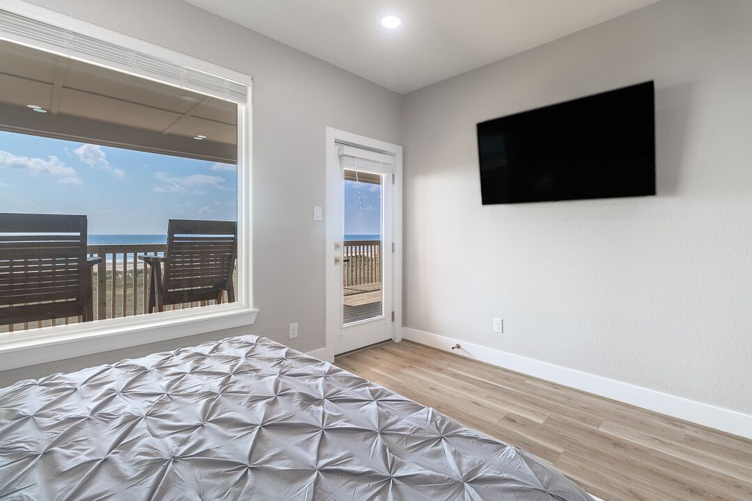 Ein Schlafzimmer mit Bett, einem an der Wand befestigten Fernseher, einem Fenster mit Blick auf den Strand und einer Tür zu einem Außenbereich.