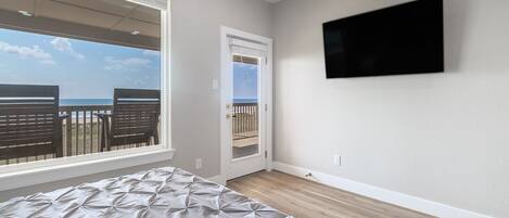 Ein Schlafzimmer mit Bett, einem an der Wand befestigten Fernseher, einem Fenster mit Blick auf den Strand und einer Tür zu einem Außenbereich.