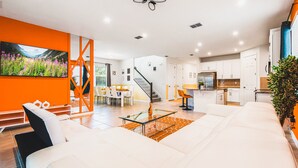 Ein modernes Wohnzimmer mit einem weißen Sofa, einem Glastisch und einem Essbereich mit gelbem Tisch und Stühlen. Es gibt eine Küche mit weißen Schränken und einem Edelstahl-Kühlschrank. Eine Treppe führt zu einer oberen Ebene.