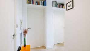 A hallway with a white door, a bookshelf filled with books, a framed picture on the wall, and a vase with dried flowers on a small orange table. - Central Location Flat near Old St and Dalston Overlooking Rosemary Park and Regents Canal - Pass the Keys (London)