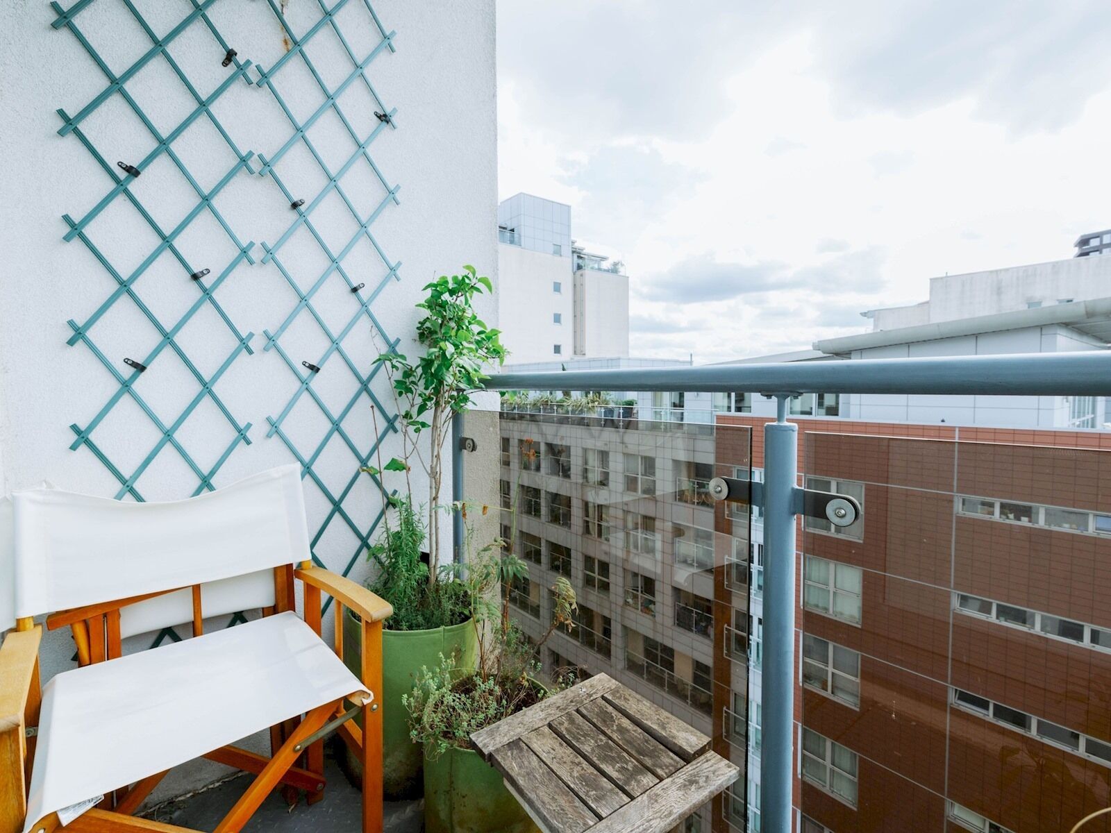 En balkon med en træstol, et lille bord, en potteplante og en væg af metalgitter.
