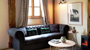 Una sala de estar acogedora con un sofá oscuro, una mesa de centro redonda, una mesita auxiliar pequeña, una chimenea y un cuadro en la pared.