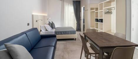 A modern hotel room with a blue sofa, a bed, a wooden dining table, and a built-in wardrobe.