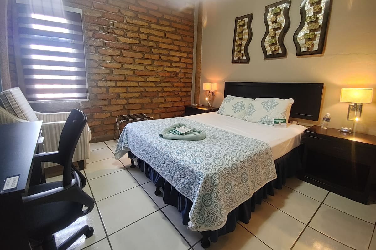 Hotelkamer met een bakstenen muur, een bed met behang, een bureau met stoel en twee bedlampjes.