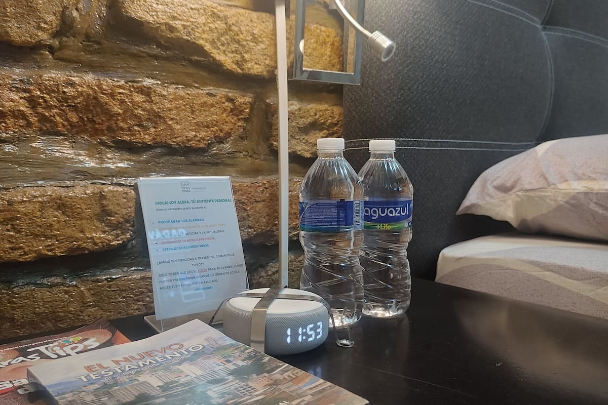 Hotelkamer met een stenen muur, een bed, nachtkastje met een klok, twee waterflessen en een boek.