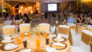 A banquet hall with round tables set for a formal event, featuring white and gold decor, and a projection screen in the background.