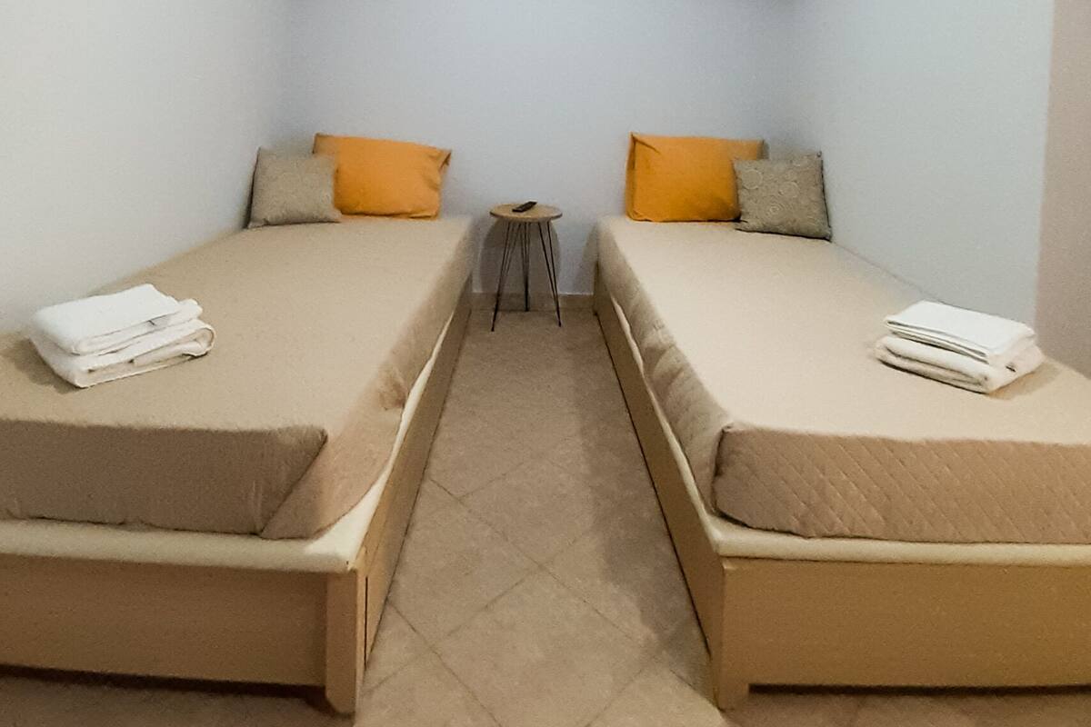 A small, narrow room with two single beds, each with a pillow and a folded towel. A small table is positioned between the beds.