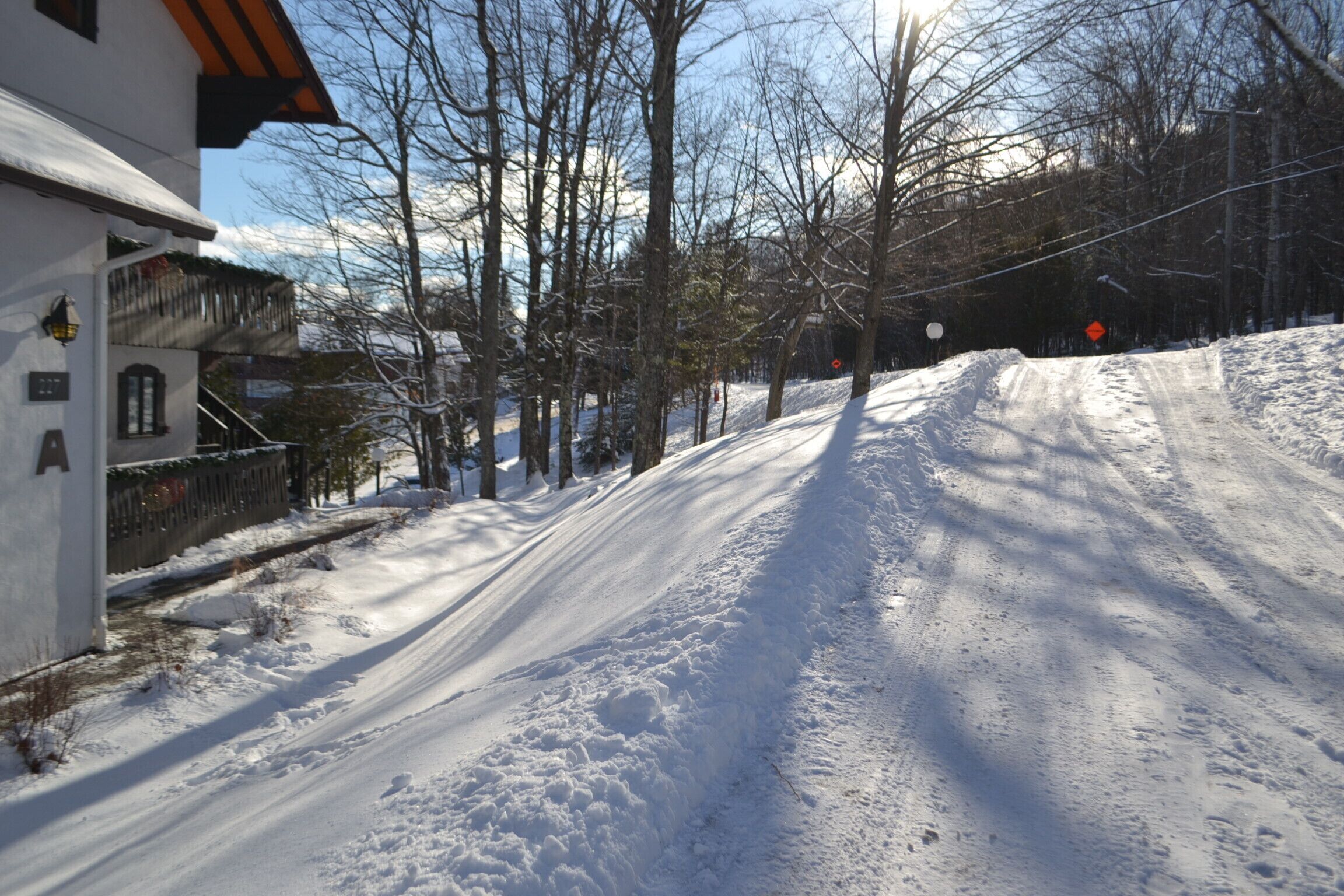 A snow-covered road with a building on the left and bare trees in the background.