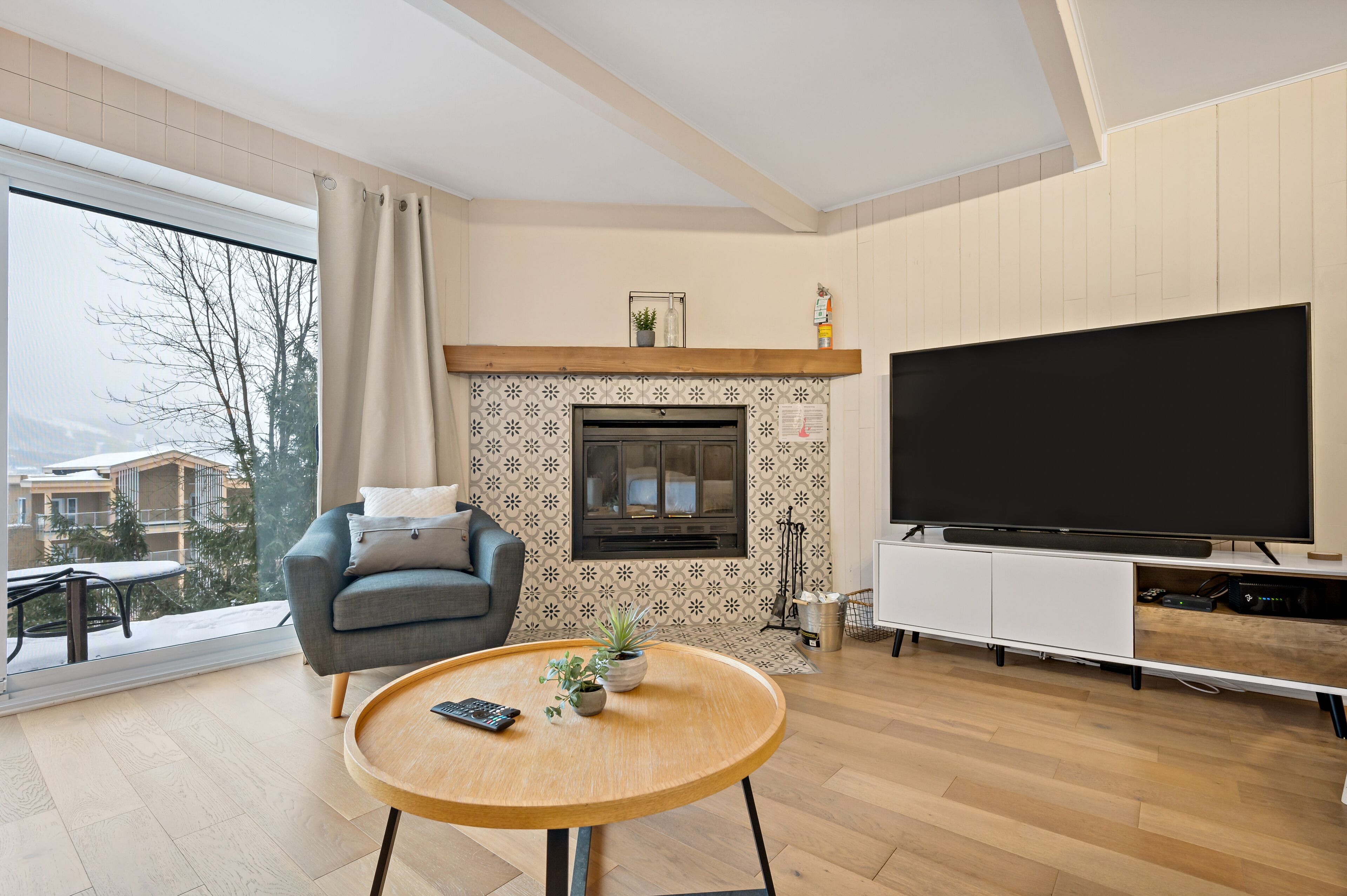 A modern living room with a fireplace, a large TV, and a balcony with a view of buildings.
