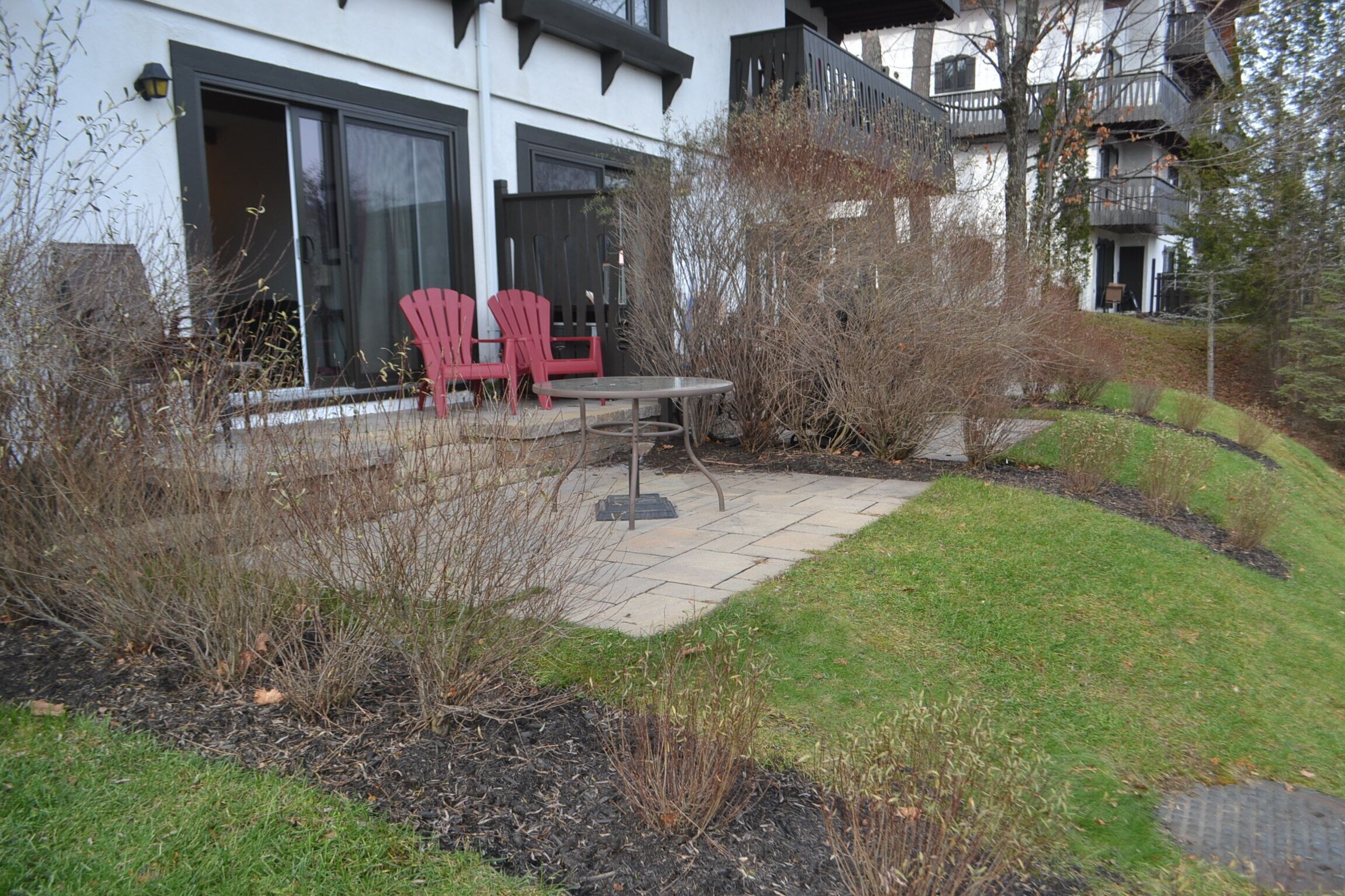 A patio with two red chairs and a table, surrounded by a landscaped garden with a grassy area and shrubs.